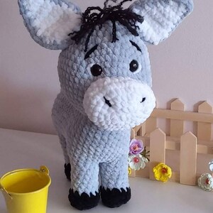 May include: A handmade, plush donkey toy in shades of gray and white. The donkey has large ears, black eyes, and a fluffy white muzzle. It has black hooves and a small tuft of black yarn for hair. A yellow bucket sits nearby.