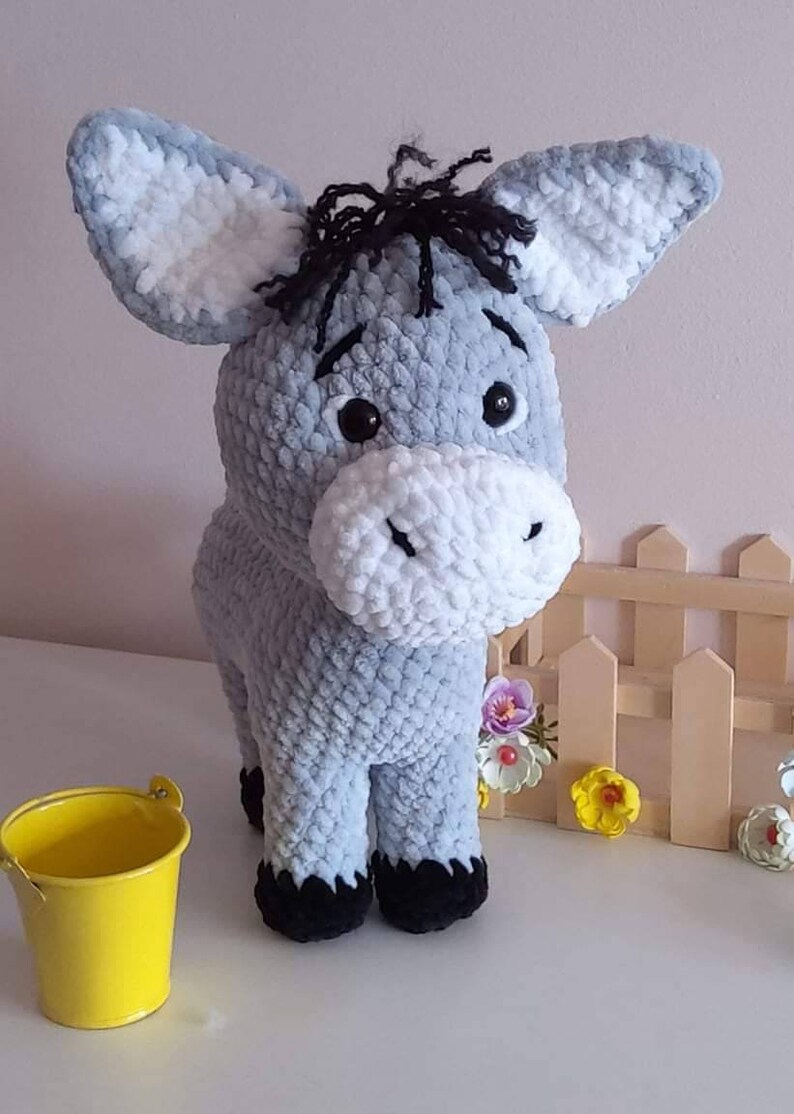 May include: A handmade, plush donkey toy in shades of gray and white. The donkey has large ears, black eyes, and a fluffy white muzzle. It has black hooves and a small tuft of black yarn for hair. A yellow bucket sits nearby.