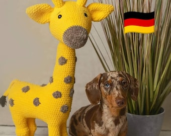 May include: A yellow crocheted giraffe toy with brown spots stands on a white wooden surface. A brown and white dachshund dog sits beside the giraffe, looking at the camera. The background features a potted plant with green leaves and two flags, the American flag and the German flag.