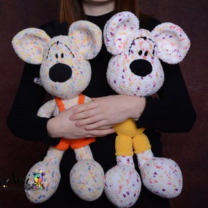 Könnte beinhalten: Zwei gehäkelte Stoffmäuse, eine mit orangefarbenen Latzhosen und die andere mit gelben Latzhosen. Beide Mäuse haben weißes Fell mit bunten Flecken und schwarzen Augen.