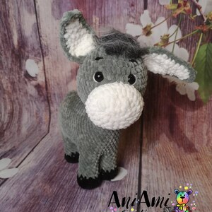 May include: A handmade gray and white crochet donkey plush toy. The donkey has large ears, a fluffy white muzzle, and black eyes. The plush animal is standing on a wooden surface. The logo "AniAmi Creations" is visible.