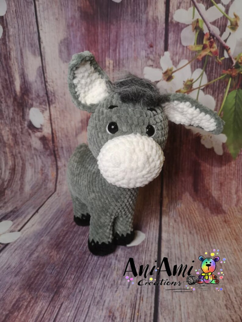May include: A handmade gray and white crochet donkey plush toy. The donkey has large ears, a fluffy white muzzle, and black eyes. The plush animal is standing on a wooden surface. The logo "AniAmi Creations" is visible.