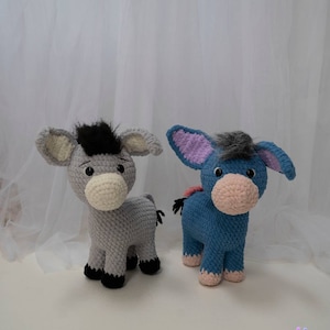 May include: Two handmade crochet donkey plushies. One is gray with a black mane, the other is blue with a gray mane. Both have large ears, white muzzles, and black hooves. The plushies are standing on a white surface.