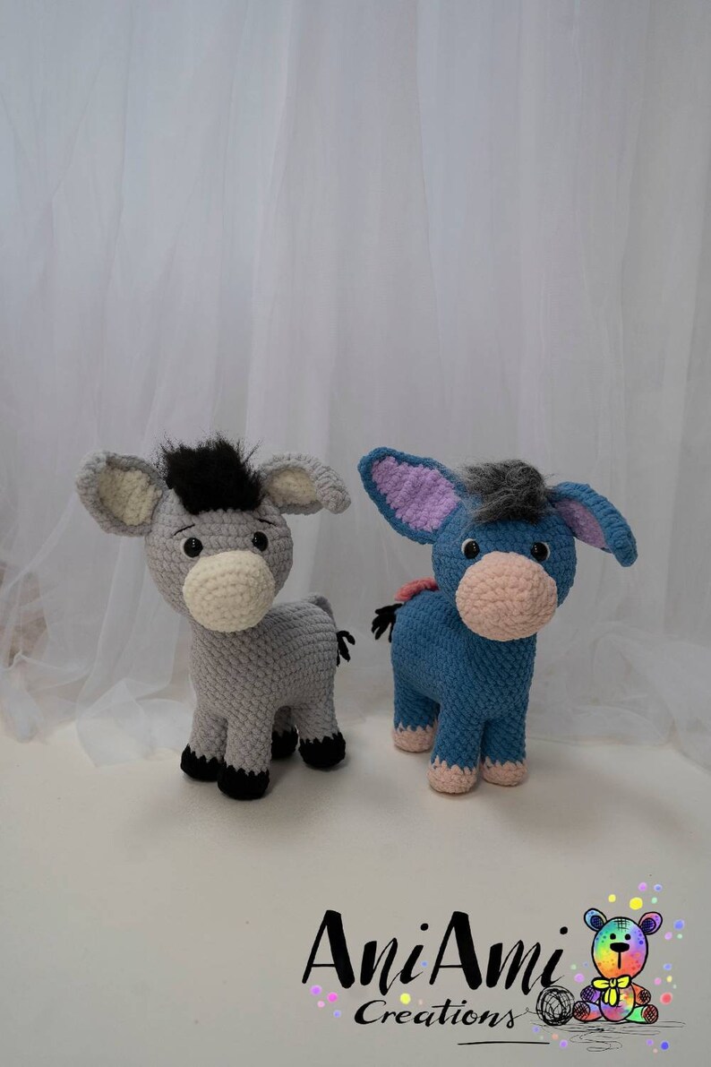 May include: Two handmade crochet donkey plushies. One is gray with a black mane, the other is blue with a gray mane. Both have large ears, white muzzles, and black hooves. The plushies are standing on a white surface.