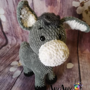 May include: A handmade gray and white plush donkey toy with large ears and a fluffy white muzzle. The donkey has black eyes and black hooves. The toy is made of soft, textured yarn. The logo "Ani Ami Creations" is visible in the bottom right corner.