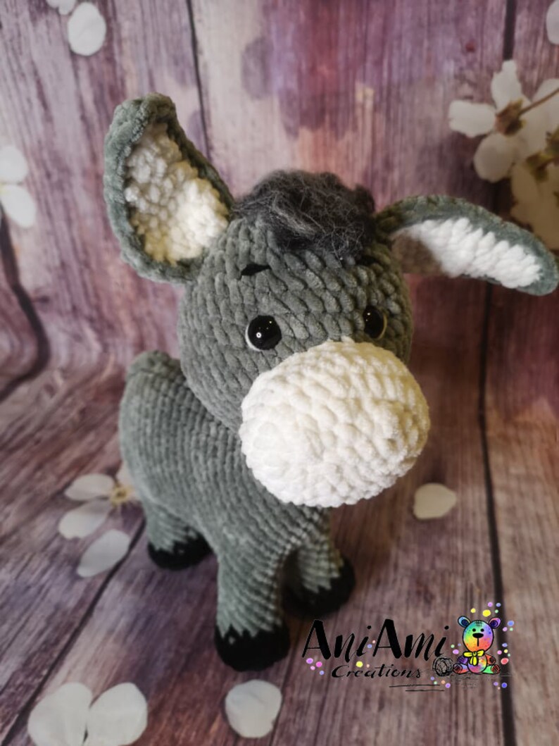 May include: A handmade gray and white plush donkey toy with large ears and a fluffy white muzzle. The donkey has black eyes and black hooves. The toy is made of soft, textured yarn. The logo "Ani Ami Creations" is visible in the bottom right corner.