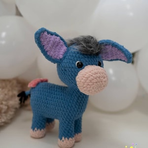 May include: A blue crochet donkey with a pink muzzle, ears, and hooves. The donkey has a gray mane and a small pink tail. The donkey is a handmade item, likely a toy or decorative item. The background includes white balloons.