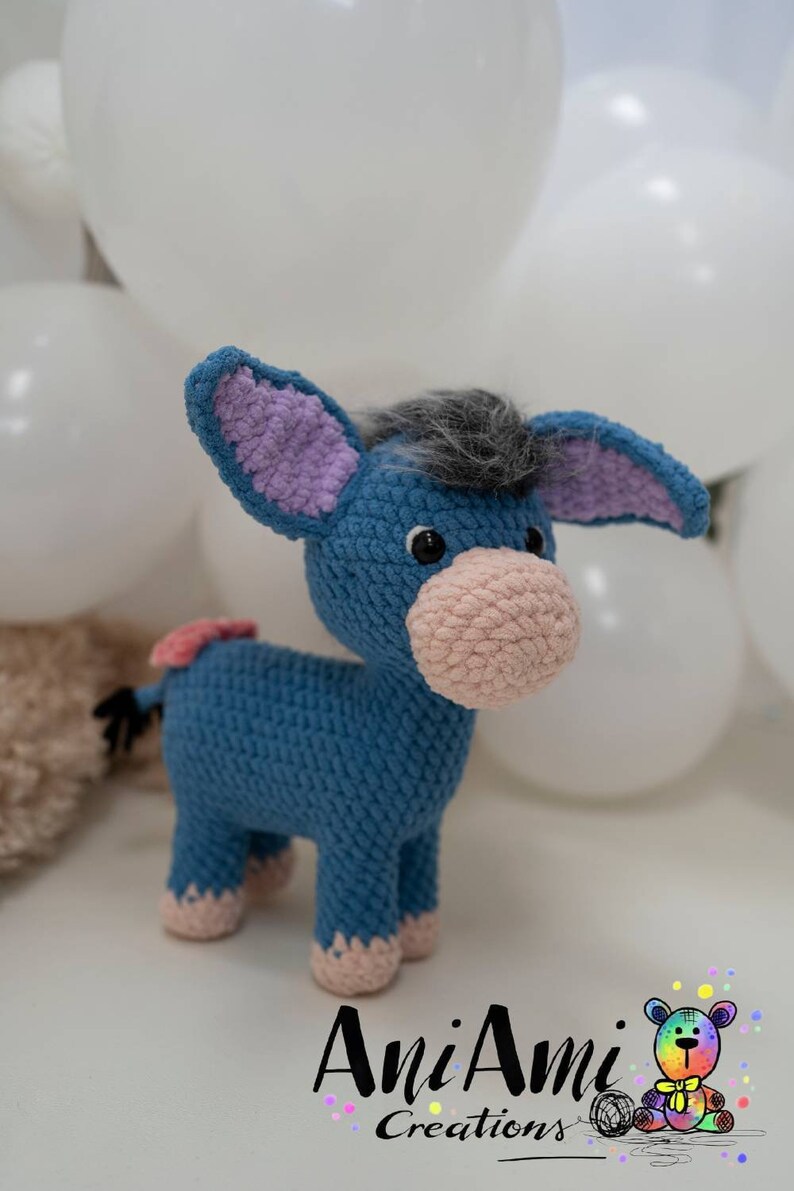 May include: A blue crochet donkey with a pink muzzle, ears, and hooves. The donkey has a gray mane and a small pink tail. The donkey is a handmade item, likely a toy or decorative item. The background includes white balloons.