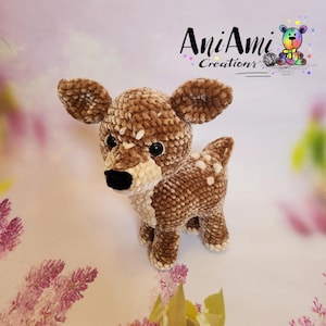 May include: A crocheted fawn stuffed animal with brown and white fur, brown eyes, and a black nose. The fawn is standing on a white background with pink and purple flowers.
