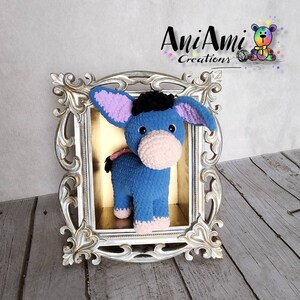 May include: A handmade blue crochet donkey toy with a pink muzzle and inner ears, black hair, and a small pink bow. The toy is displayed in an ornate silver frame with the text "AniAmi Creations".