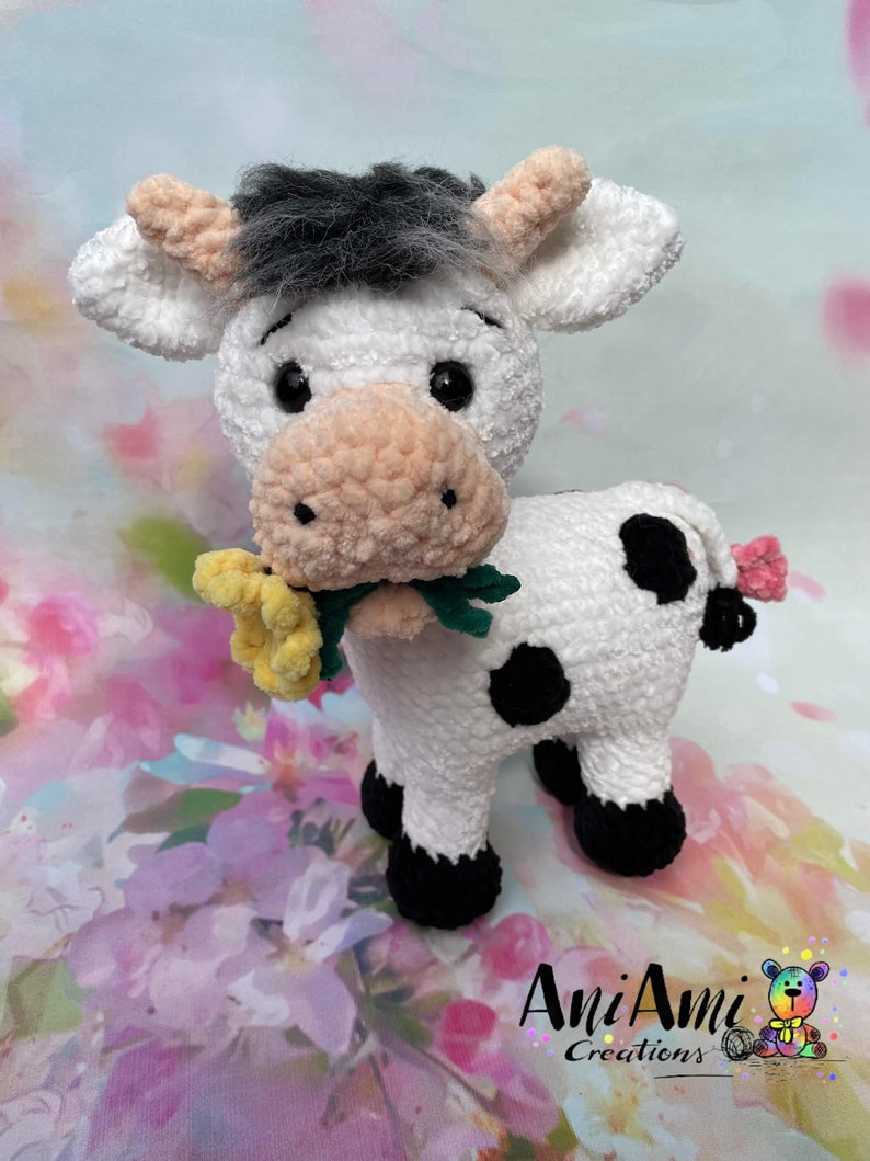 Puede incluir: Un juguete de peluche de vaca blanco tejido a crochet con manchas negras, sosteniendo una flor amarilla en su boca. La vaca tiene una cara blanca y negra con ojos negros y una nariz rosa. El juguete est&aacute; de pie sobre un fondo floral.