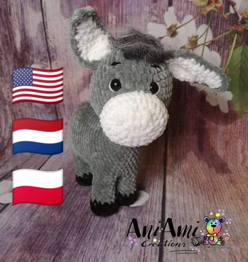 May include: A handmade gray plush donkey with a white muzzle and inner ears. The donkey has black eyes and hooves. The plush animal is standing on a wooden surface. The image includes the American flag, the Dutch flag, and the Polish flag.