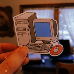 May include: A sticker featuring a cartoon illustration of a vintage computer setup. The design includes a beige tower, a monitor with a blue screen, a keyboard, and a mouse with an orange mousepad. The sticker is held by a hand.