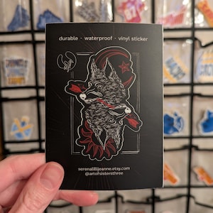 May include: Black and white illustration of a wolf's head with a red crescent moon and star. The sticker is on a black background with the text "durable - waterproof - vinyl sticker" and the artist's website and social media handle.