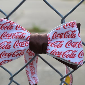 May include: A white bow tie with a brown center, featuring a repeated pattern of the Coca-Cola logo in red.