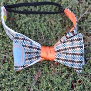 May include: A plaid bow tie with blue, black, and orange accents. The bow tie has a light blue patch with orange stitching. The center is a solid orange color, and the adjustable strap is black.