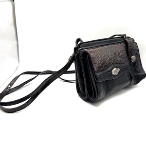 May include: Black crossbody bag with a brown, textured flap and strap. The bag features a silver-toned emblem on the front and a long, adjustable strap. The bag has a textured, faux-leather appearance.