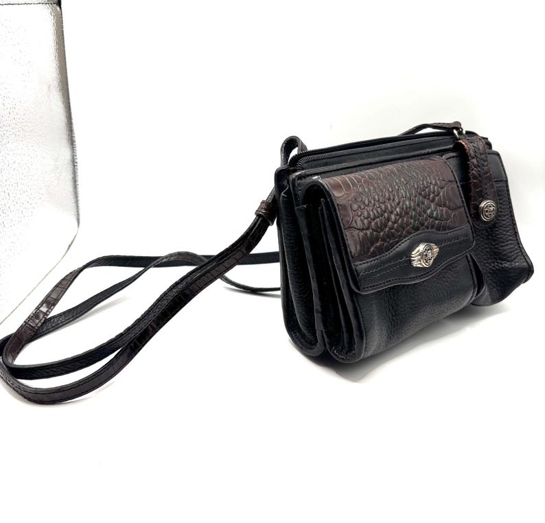 May include: Black crossbody bag with a brown, textured flap and strap. The bag features a silver-toned emblem on the front and a long, adjustable strap. The bag has a textured, faux-leather appearance.