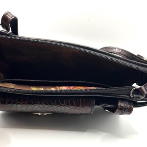 May include: Open view of a brown and black handbag with a textured, crocodile-like pattern. The interior reveals a dark lining and multiple compartments, including a zippered section and a small pocket. The bag has a shoulder strap and a small handle.