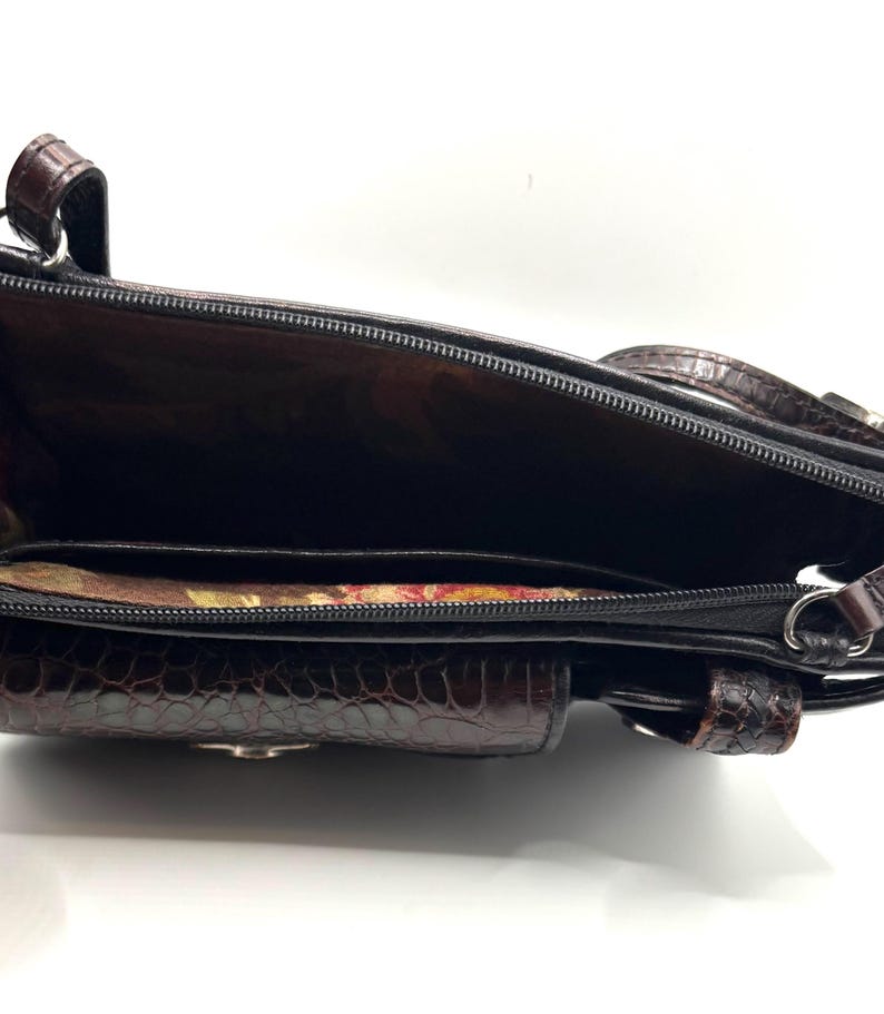May include: Open view of a brown and black handbag with a textured, crocodile-like pattern. The interior reveals a dark lining and multiple compartments, including a zippered section and a small pocket. The bag has a shoulder strap and a small handle.