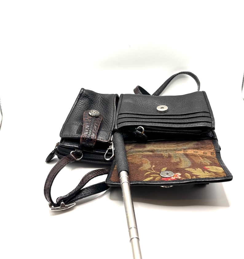 May include: Black leather crossbody bag with multiple compartments. Features a textured exterior, a decorative clasp, and a brown strap. The interior lining has a floral pattern. Includes a silver selfie stick.