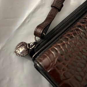 May include: A brown and black wallet with a wrist strap. The wallet has a textured, crocodile-like pattern on the front. A silver heart-shaped charm hangs from the zipper pull.