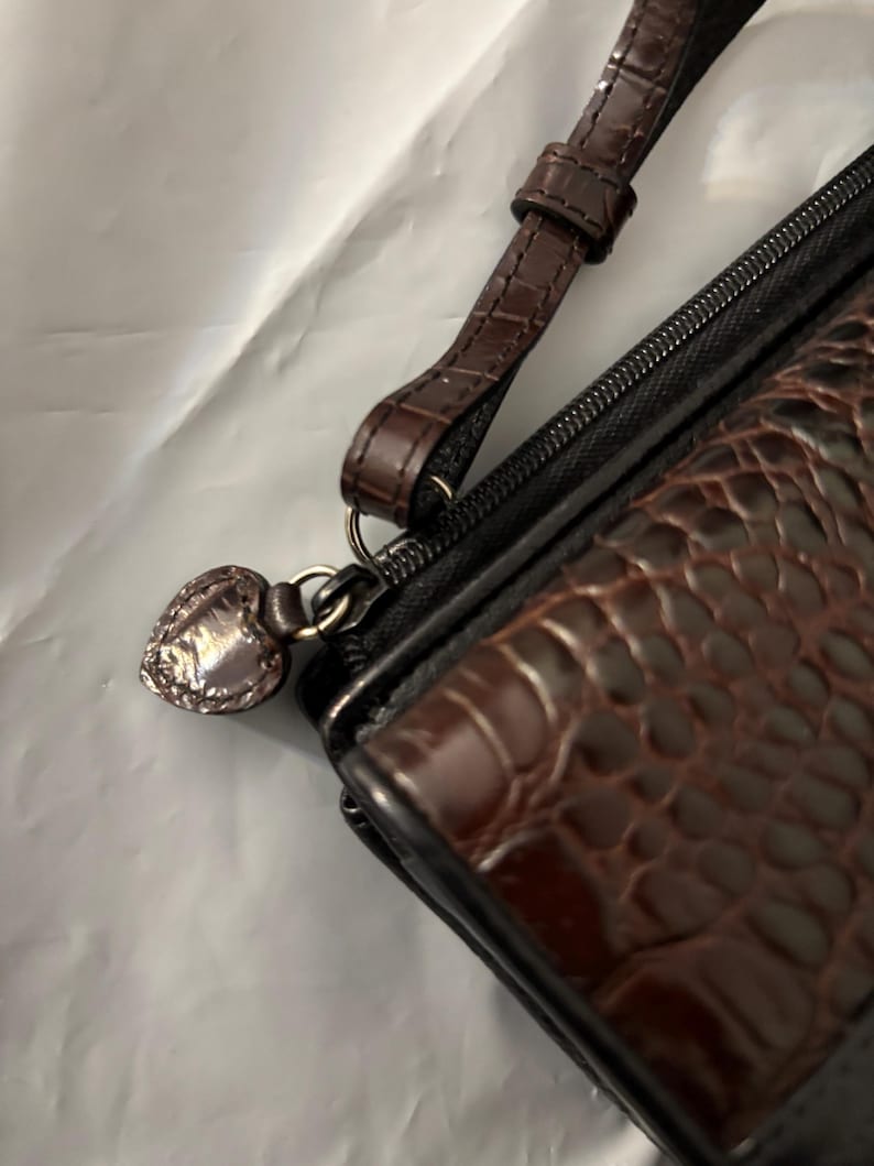 May include: A brown and black wallet with a wrist strap. The wallet has a textured, crocodile-like pattern on the front. A silver heart-shaped charm hangs from the zipper pull.