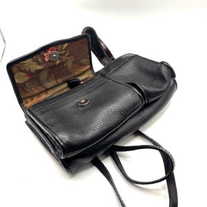 May include: Black leather crossbody bag with a textured finish. The bag features a flap closure with a silver-tone clasp and a floral patterned interior lining. The adjustable strap is visible, and the bag is open, revealing the interior.