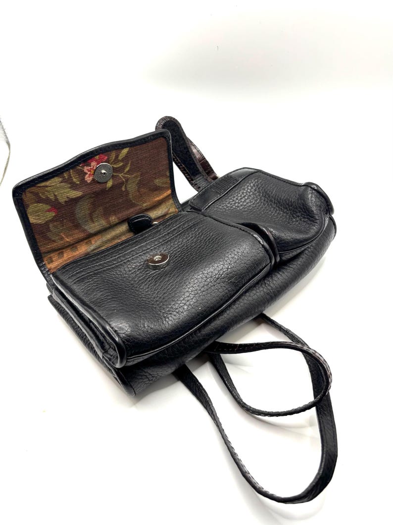 May include: Black leather crossbody bag with a textured finish. The bag features a flap closure with a silver-tone clasp and a floral patterned interior lining. The adjustable strap is visible, and the bag is open, revealing the interior.