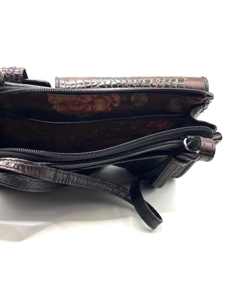 May include: Black and brown leather crossbody bag with a crocodile-like texture. The bag has a floral patterned interior and a long strap. The bag is open, revealing multiple compartments and a zipper closure.