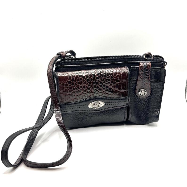 May include: Black crossbody bag with a brown, faux-crocodile skin flap and accents. The bag has a long, adjustable strap and multiple compartments. Silver-toned hardware accents the bag's design.