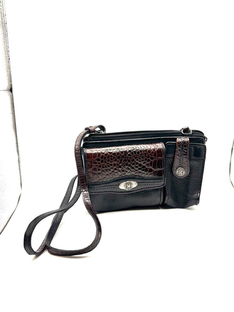 May include: Black crossbody bag with a brown, textured front pocket and a silver-tone clasp. The bag features a long, adjustable strap and a zippered main compartment. The brown pocket has a crocodile-like pattern.