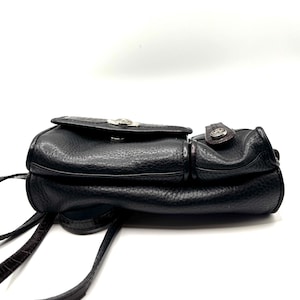 May include: Black leather crossbody bag with a textured finish. The bag features a flap closure with a silver-toned emblem and a brown strap. The bag has a small pocket on the side and a long, thin strap for carrying.