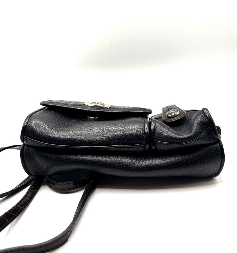 May include: Black leather crossbody bag with a textured finish. The bag features a flap closure with a silver-toned emblem and a brown strap. The bag has a small pocket on the side and a long, thin strap for carrying.
