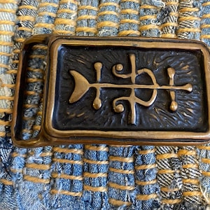 May include: A rectangular belt buckle with a dark, textured center and a bronze-colored frame. The center features an abstract design with a fish-like shape and other stylized elements. The buckle is set against a woven blue and yellow background.