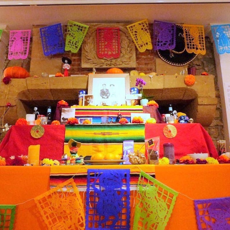 May include: A Day of the Dead altar with colorful decorations, including paper flowers, candles, and a framed photo. The altar is decorated with a variety of colorful paper cutouts, including skulls, flowers, and other traditional Day of the Dead imagery.