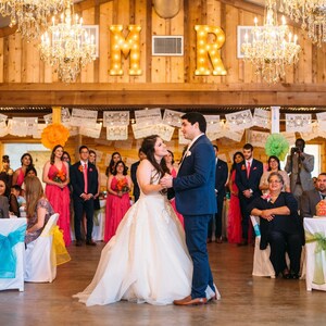 Puede incluir: Una novia y un novio bailando en su recepci&oacute;n de boda. La novia lleva un vestido de novia blanco y el novio lleva un traje azul. La recepci&oacute;n est&aacute; decorada con pancartas de papel de colores y flores. Las letras "M" y "R" est&aacute;n iluminadas detr&aacute;s de la pareja.