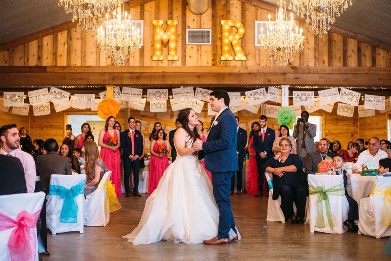 Puede incluir: Una novia y un novio bailando en su recepci&oacute;n de boda. La novia lleva un vestido de novia blanco y el novio lleva un traje azul. La recepci&oacute;n est&aacute; decorada con pancartas de papel de colores y flores. Las letras "M" y "R" est&aacute;n iluminadas detr&aacute;s de la pareja.