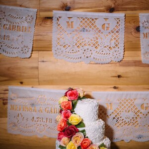 Puede incluir: Un pastel de bodas blanco de tres niveles decorado con glaseado blanco y un delicado dise&ntilde;o de espiral. El pastel est&aacute; adornado con una guirnalda de rosas rojas, naranjas y amarillas en cascada.
