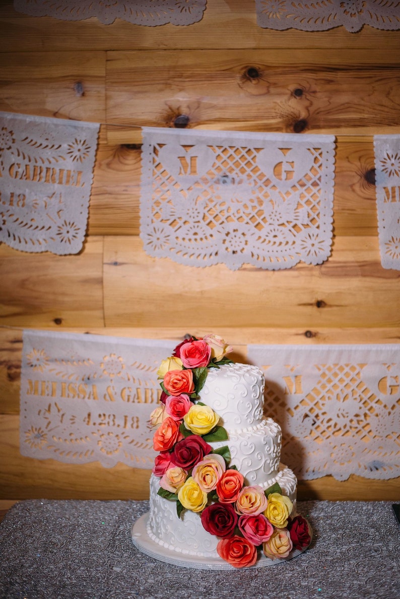 Puede incluir: Un pastel de bodas blanco de tres niveles decorado con glaseado blanco y un delicado dise&ntilde;o de espiral. El pastel est&aacute; adornado con una guirnalda de rosas rojas, naranjas y amarillas en cascada.