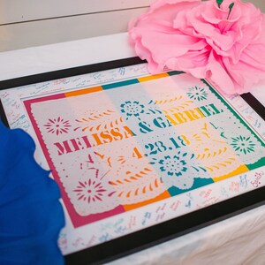 Puede incluir: Un recorte de papel blanco enmarcado con rayas de colores y el texto "Melissa & Gabriel 4.23.18" en un marco negro. El marco est&aacute; sobre una mesa blanca con una flor de papel azul y rosa a cada lado.