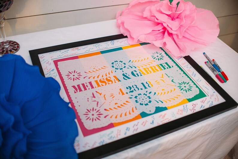 Puede incluir: Un recorte de papel blanco enmarcado con rayas de colores y el texto "Melissa & Gabriel 4.23.18" en un marco negro. El marco est&aacute; sobre una mesa blanca con una flor de papel azul y rosa a cada lado.
