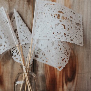 Puede incluir: Banderas de papel recortado blanco con el texto "HANNAH & GABRIEL" sobre un fondo de madera. Las banderas est&aacute;n sujetas a palos de madera y colocadas en una botella de vidrio transparente.
