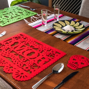 May include: A table setting with a red and green felt placemat with a cut-out design. The placemats feature the words "Amor" and "Home". There are also two skull-shaped coasters and a bottle of tequila.