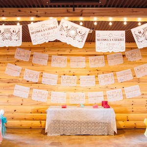 May include: A wedding reception venue decorated with white paper cutouts, or "papel picado", with the names "Melissa & Gabriel" and the date "1.28.18". The venue has a wooden wall and a table with a white tablecloth.