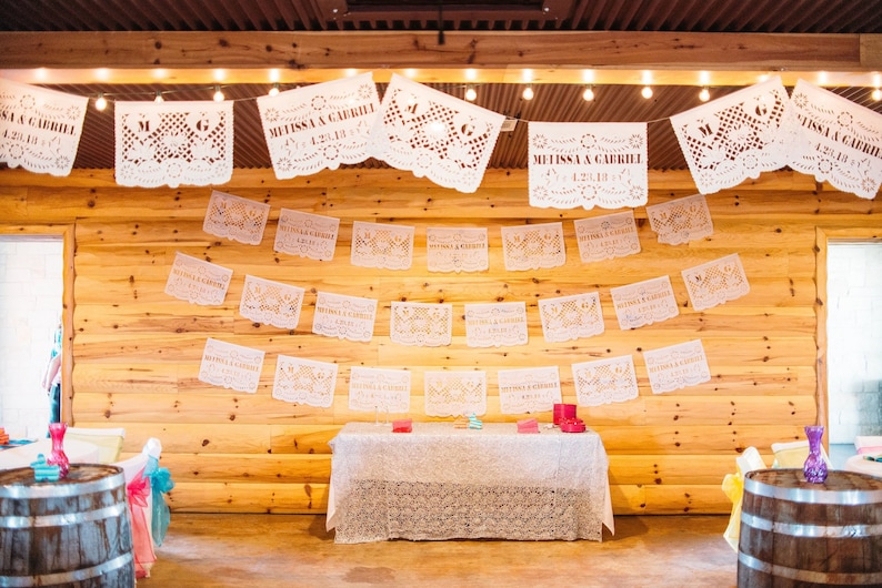 Puede incluir: Un lugar de recepci&oacute;n de boda decorado con recortes de papel blanco, o "papel picado", con los nombres "Melissa & Gabriel" y la fecha "1.28.18". El lugar tiene una pared de madera y una mesa con un mantel blanco.