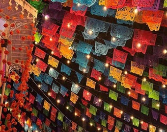 Può includere: Diverse file di bandierine rettangolari in carta colorata con intricati motivi ritagliati sono appese in uno spazio all'aperto durante la notte. Le bandierine, nei toni del rosso, arancione, giallo, verde, blu e viola, sono illuminate da luci.
