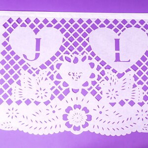 Puede incluir: Bandera de papel blanco con un dise&ntilde;o decorativo de corte. La bandera tiene dos corazones con las letras "J" y "L" cortadas. La bandera tambi&eacute;n tiene un dise&ntilde;o floral con la palabra "cruz" cortada.