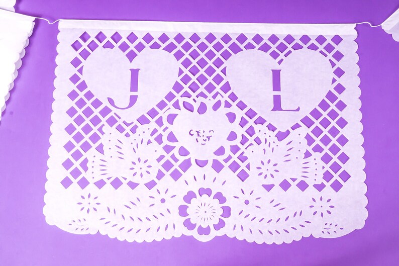 Puede incluir: Bandera de papel blanco con un dise&ntilde;o decorativo de corte. La bandera tiene dos corazones con las letras "J" y "L" cortadas. La bandera tambi&eacute;n tiene un dise&ntilde;o floral con la palabra "cruz" cortada.