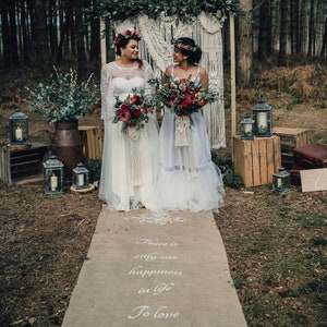 May include: Two people wearing white wedding dresses stand in a forest with a macrame backdrop. They are holding bouquets of red flowers. The aisle is decorated with a burlap runner with the text "There is only one happiness in life To love and be loved".
